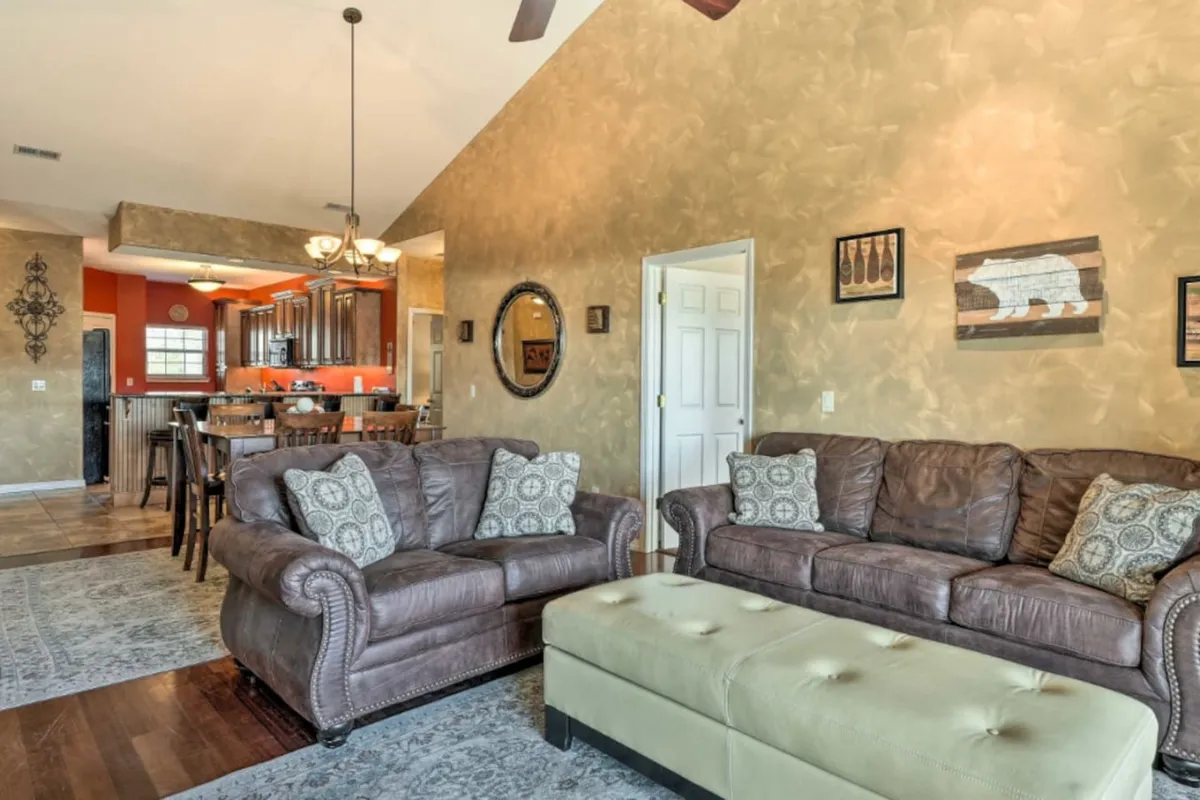 A stylish, cozy living area featuring leather sofas, textured walls, and decorative pillows, with a round mirror accenting the space. The kitchen area can be seen in the background, underlining the seamless transition between lounging and dining spaces. The overall mood is warm and inviting, fostering both casual relaxation and lively gatherings.