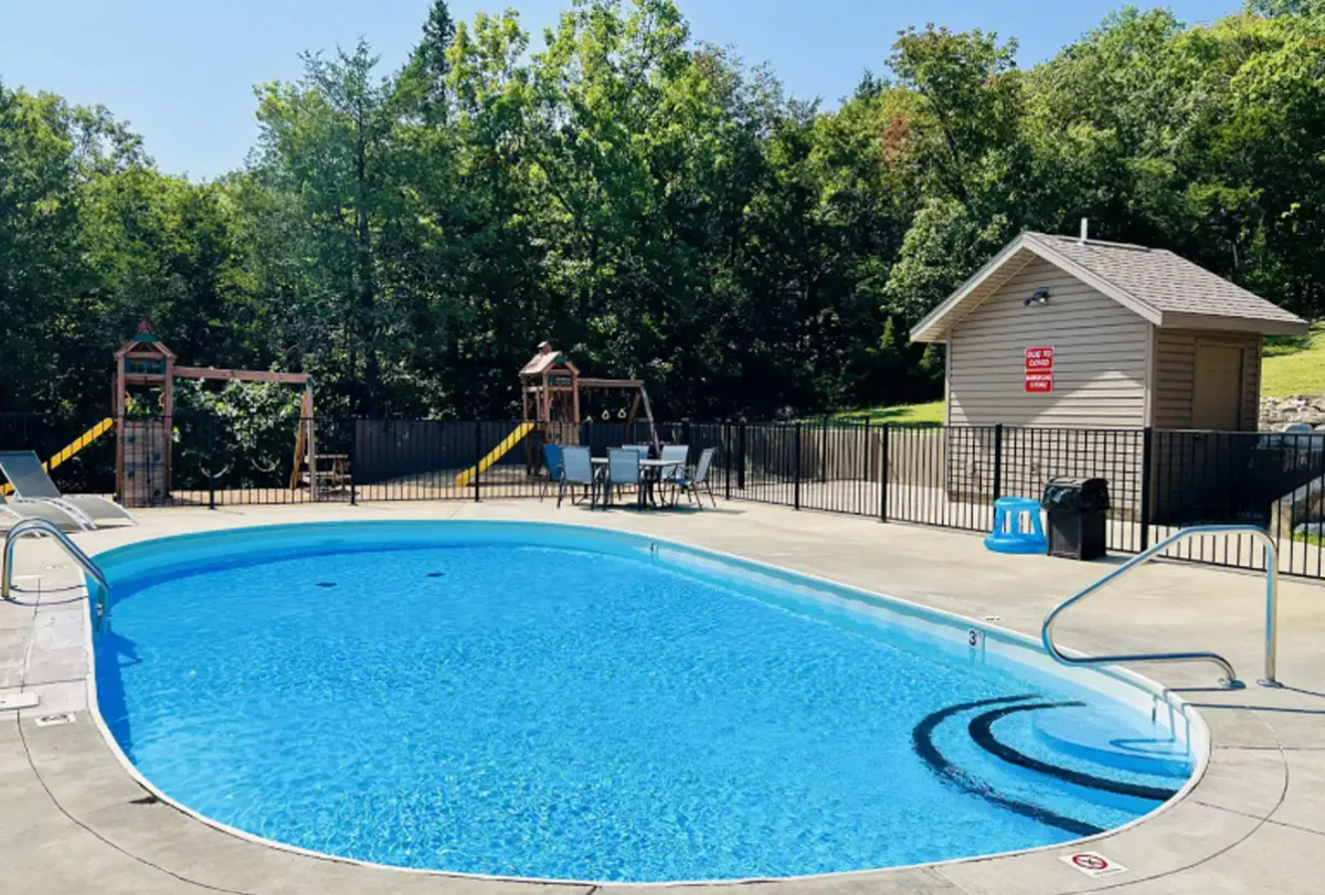 This exterior image features a lively community swimming pool area with people swimming, sunbathing, and children playing on playground equipment nearby. Outdoor seating and lush greenery surround the pool, creating a vibrant, family-friendly atmosphere.
