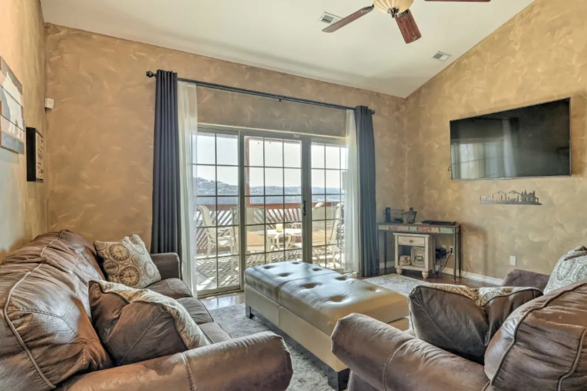 A broad, sun-filled living room with a large sectional sofa arranged around an ottoman. Sliding glass doors let in abundant natural light, and a television is visible, creating a hub for conversation, relaxation, and entertainment. The seating area offers a view out to the surrounding landscape, reinforcing the 'open-concept' and 'togetherness' theme.