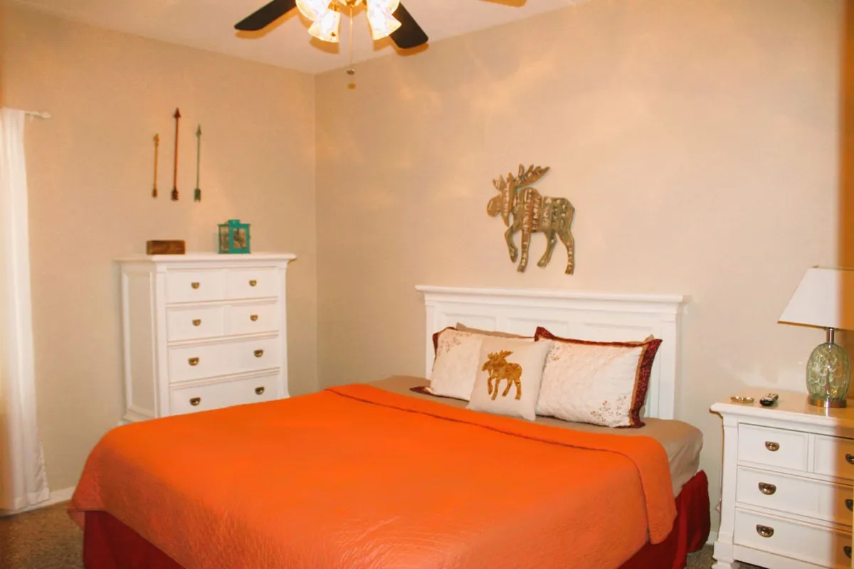 A cozy bedroom with a bed topped by an orange coverlet and a decorative moose pillow, complemented by white dressers and playful wall art. The space feels warm and inviting, ideal for recharging after a busy day.