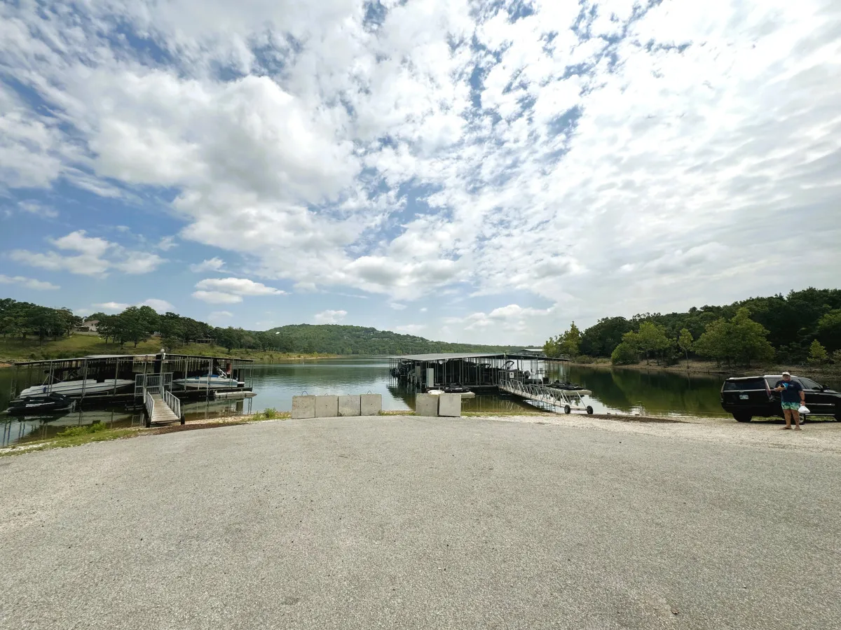 A lakeside dock extending into calm waters with rolling hills in the background, a few vehicles and clouds overhead. The scene conveys easy lake access for boating, fishing, or simply enjoying the outdoors.